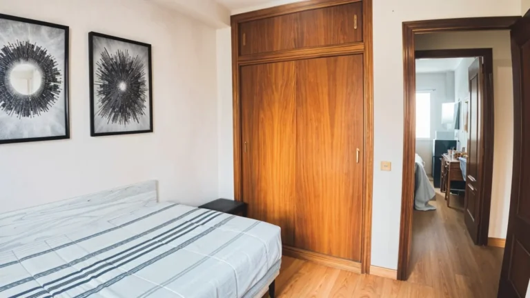 Un dormitorio con una cama, ropa de cama a rayas, dos obras de arte abstracto en la pared, un armario de madera y una puerta abierta que da a otra habitación.