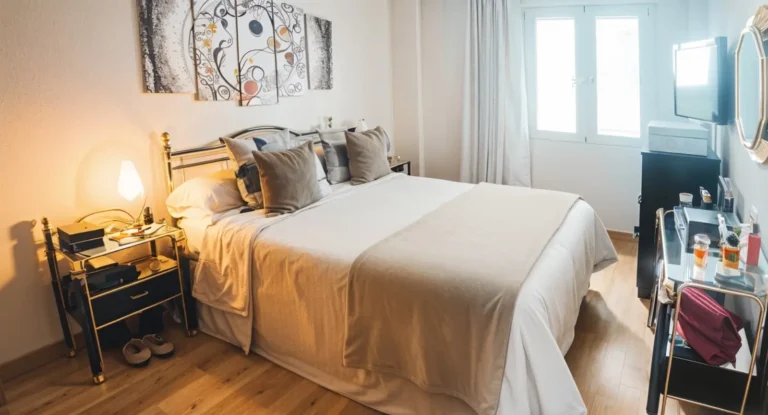 Una cama doble bien hecha con ropa de cama beige, almohadas y mesillas de noche en un dormitorio luminoso con suelos de madera y una ventana que deja entrar la luz natural.
