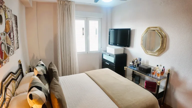 Una cama bien hecha frente a un televisor montado en la pared; una cómoda, un carrito de bebidas y un espejo redondo están a la derecha, con una ventana que deja entrar la luz natural.