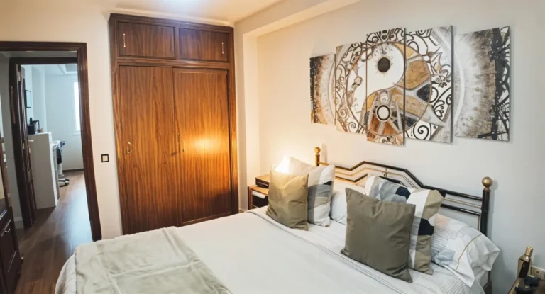 En un dormitorio bien iluminado, junto a un armario empotrado de madera, hay una cama de matrimonio bien hecha con varias almohadas y una pared decorada con arte abstracto.