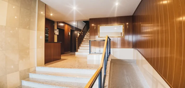 Vista interior de un edificio moderno con paredes de azulejos beige y paneles de madera, una escalera con barandilla y un gran espejo en la pared.