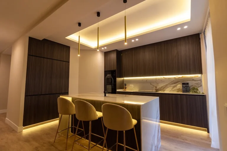 Cocina moderna con armarios de madera oscura, isla de mármol, taburetes de bar beige y cálida iluminación empotrada y colgante.