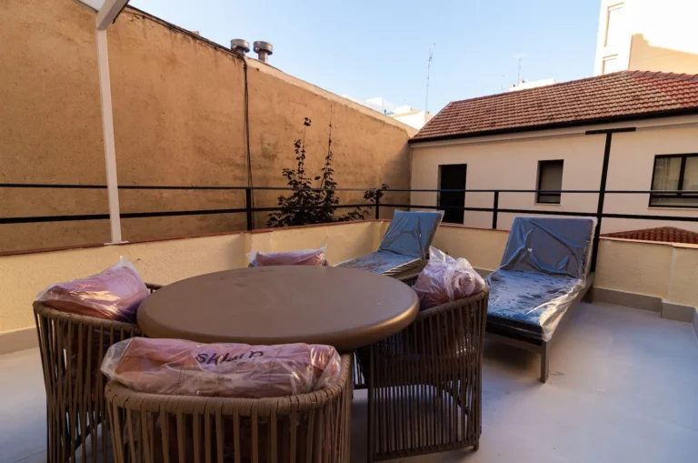Terraza exterior con una mesa redonda, cuatro sillas y dos tumbonas, todas con fundas protectoras de plástico, contra una pared beige y el edificio adyacente.