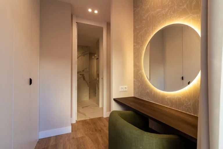 Una habitación moderna con suelo de madera, una silla verde en un escritorio de madera oscura, un espejo redondo retroiluminado en la pared y una puerta abierta que revela un cuarto de baño de mármol.