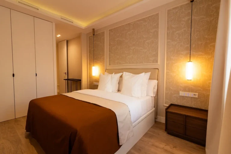 Una cama bien hecha con sábanas blancas y una manta marrón en un dormitorio moderno, con lámparas colgantes, una mesilla de noche de madera y grandes armarios empotrados.