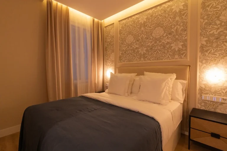 Una cama bien hecha con almohadas blancas y una manta gris oscuro en un dormitorio suavemente iluminado con papel pintado estampado y cortinas beige.