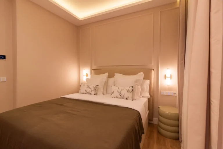 Un dormitorio moderno con paredes de color beige, una cama de matrimonio con almohadas blancas y ropa de cama marrón, dos lámparas de pared y una mesa auxiliar redonda de color verde.