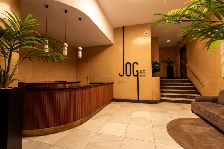 Vestíbulo de un edificio moderno con mostrador de recepción de madera, paredes y suelo de baldosas beige, plantas en macetas y escalones que conducen a un pasillo; en la pared se lee "JOG 65".