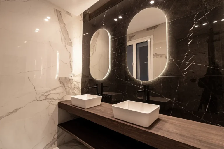 Moderno cuarto de baño con dos lavabos rectangulares sobre encimera de madera, pared de mármol negro con espejos ovalados e iluminación empotrada.