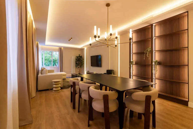 Comedor y sala de estar modernos con una larga mesa negra, sillas acolchadas, un sofá blanco, TV montada en la pared, estanterías empotradas, lámpara de araña y una gran ventana con cortinas.