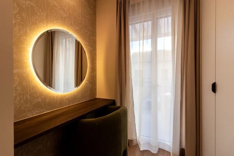 Un moderno tocador con un espejo redondo retroiluminado, encimera de madera oscura, silla verde y cortinas transparentes que cubren una ventana por la que entra luz natural.