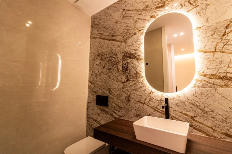 Moderno cuarto de baño con paredes de mármol, un espejo oval retroiluminado, un lavabo rectangular sobre un tocador de madera e iluminación empotrada que se refleja en las superficies brillantes.
