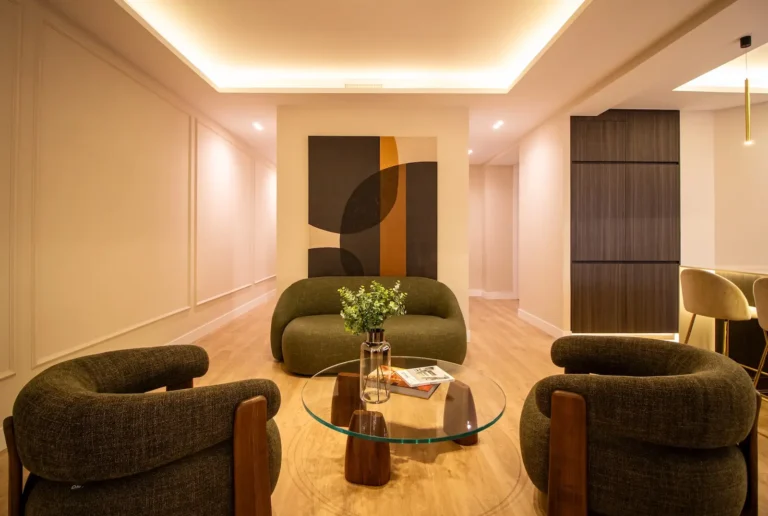 Moderno salón con sillones verde oscuro, una mesa de centro de cristal con un jarrón de flores, arte abstracto en la pared y una cálida iluminación empotrada en el techo.