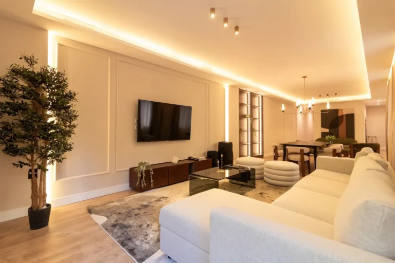Moderno salón con un gran sofá seccional beige, televisor montado en la pared, mesas de centro de cristal, iluminación contemporánea, planta de interior y zona de comedor en una distribución diáfana.