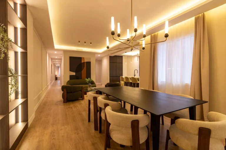 Moderna zona de comedor y sala de estar con una larga mesa negra, sillas acolchadas de color beige, lámpara de araña, suelo de madera e iluminación tenue. Estanterías y grandes ventanales a la vista, junto con una decoración contemporánea.