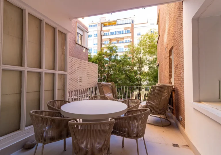 Un balcón cubierto con una mesa redonda y seis sillas ofrece vistas a los árboles y a los edificios de apartamentos del fondo.