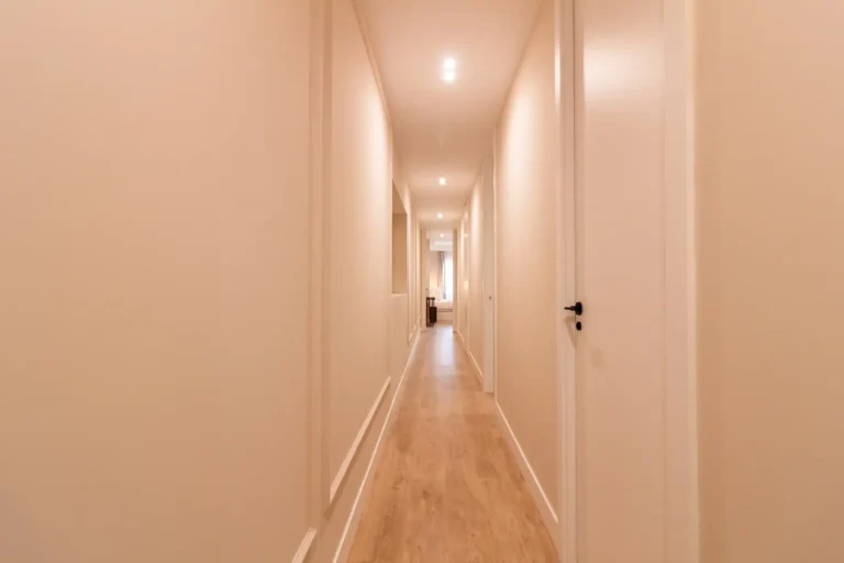 Un pasillo largo y estrecho con paredes claras, suelo de madera, luces empotradas en el techo y varias puertas a ambos lados que conducen a una habitación al final.