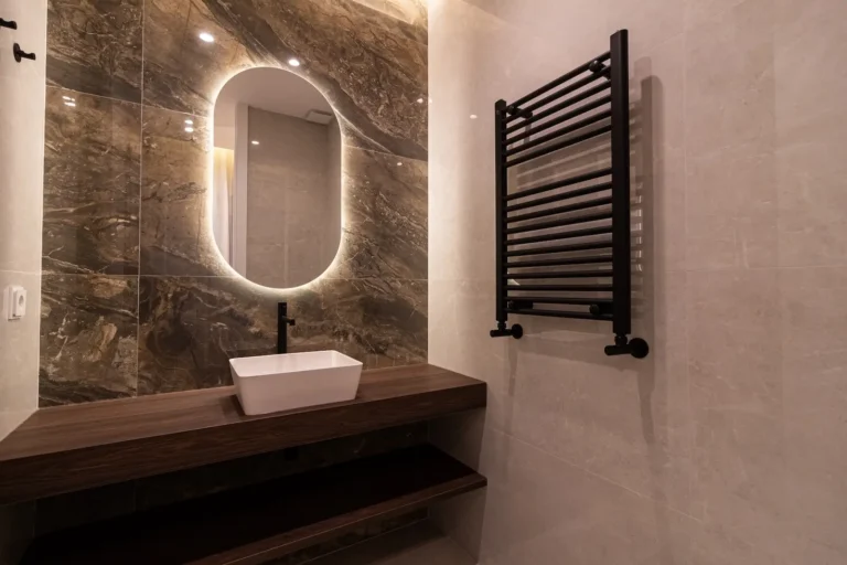 Moderno cuarto de baño con lavabo rectangular, encimera de madera, espejo oval retroiluminado, pared de mármol y toallero calefactor negro.